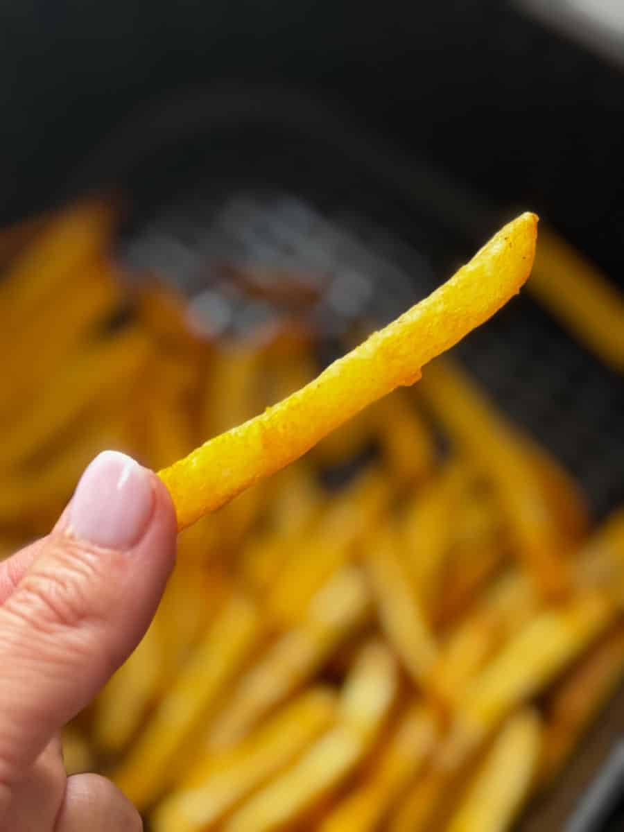 Hand holding a crispy French fry with a tray of fries in the background.