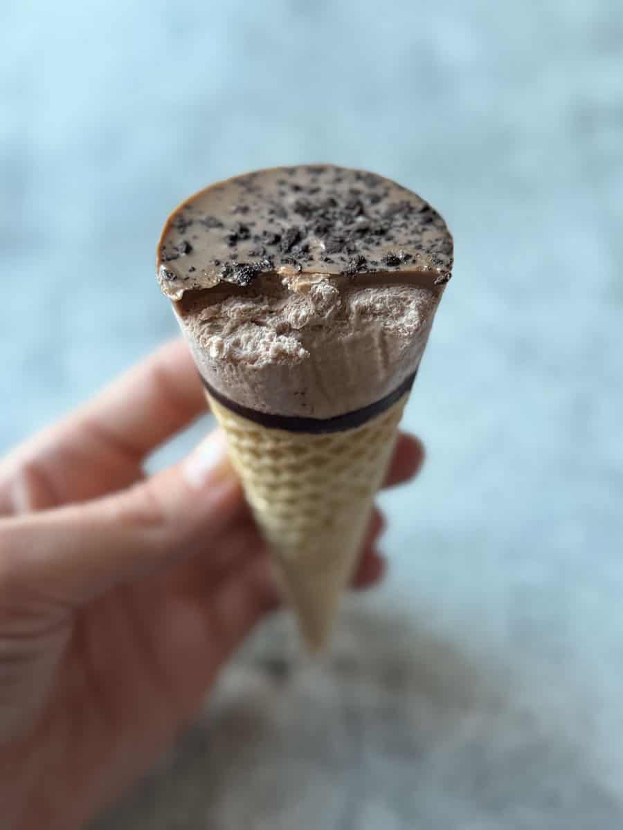 Ice cream cone with cookies and cream flavor, topped with cookie crumbles, held in hand against a ne.