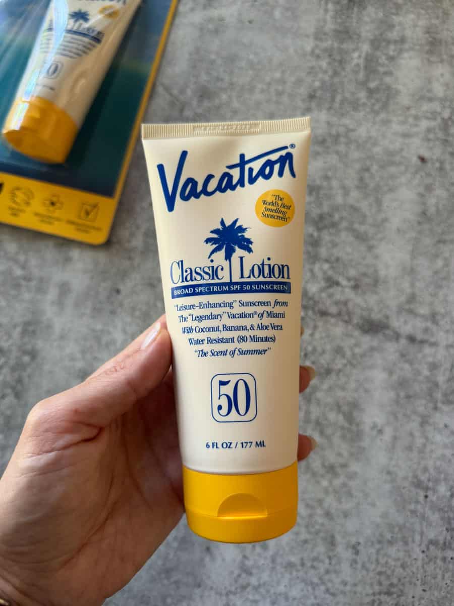 Hand holding Vacation Classic Lotion SPF 50 sunscreen tube with beach background.