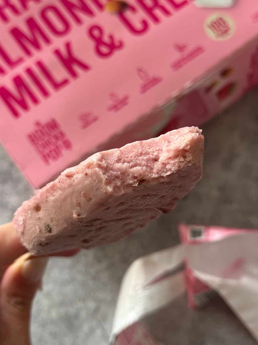 Close-up of a creamy lemon and milk cheesecake bar held in hand, with pink cheesecake box in backgro.