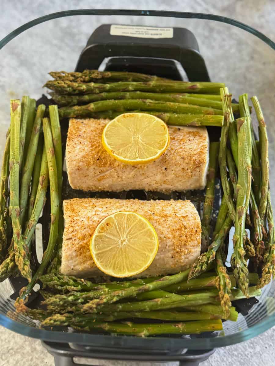 Baked fish fillets with lemon slices, surrounded by roasted asparagus spears in a glass dish.